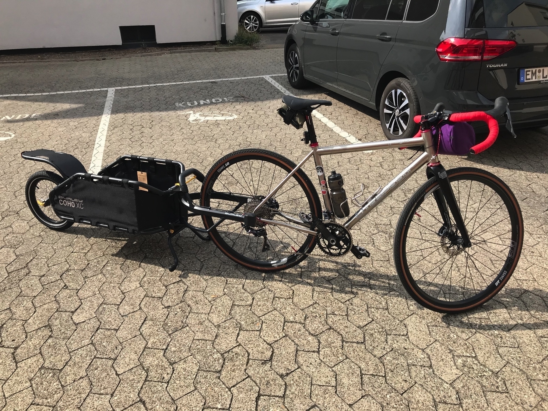 Drop bar, titanium gravel bike with a single-wheel cargo trailer attached to the rear axle in a parking lot.