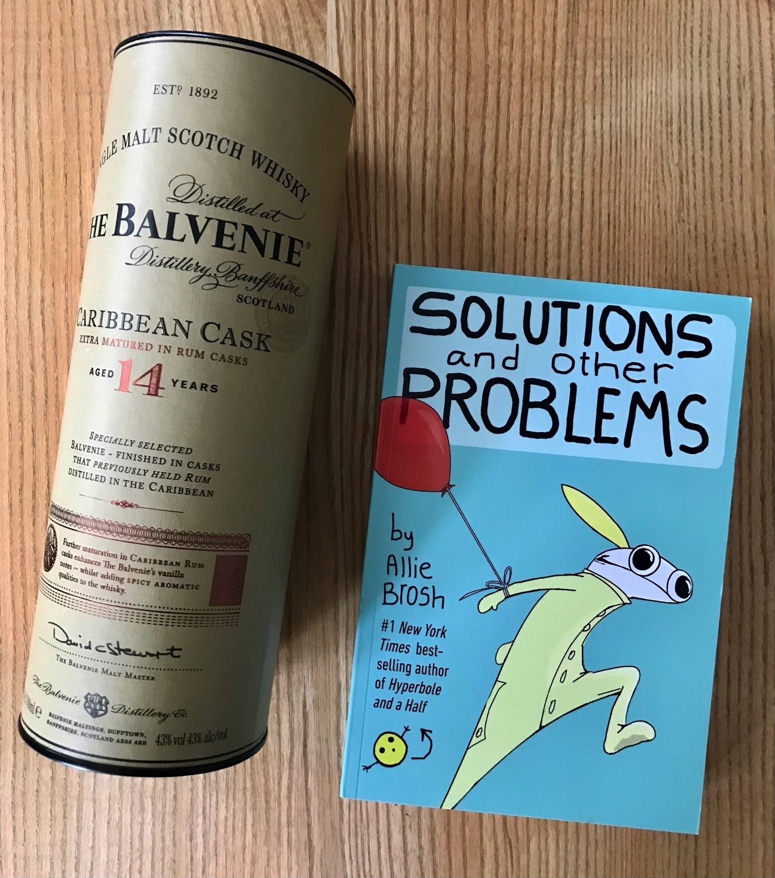 Tubublar packaging containing a bottle of Balvenie Caribbean Cask whisky and the new book by Allie Brosh on a wooden table.