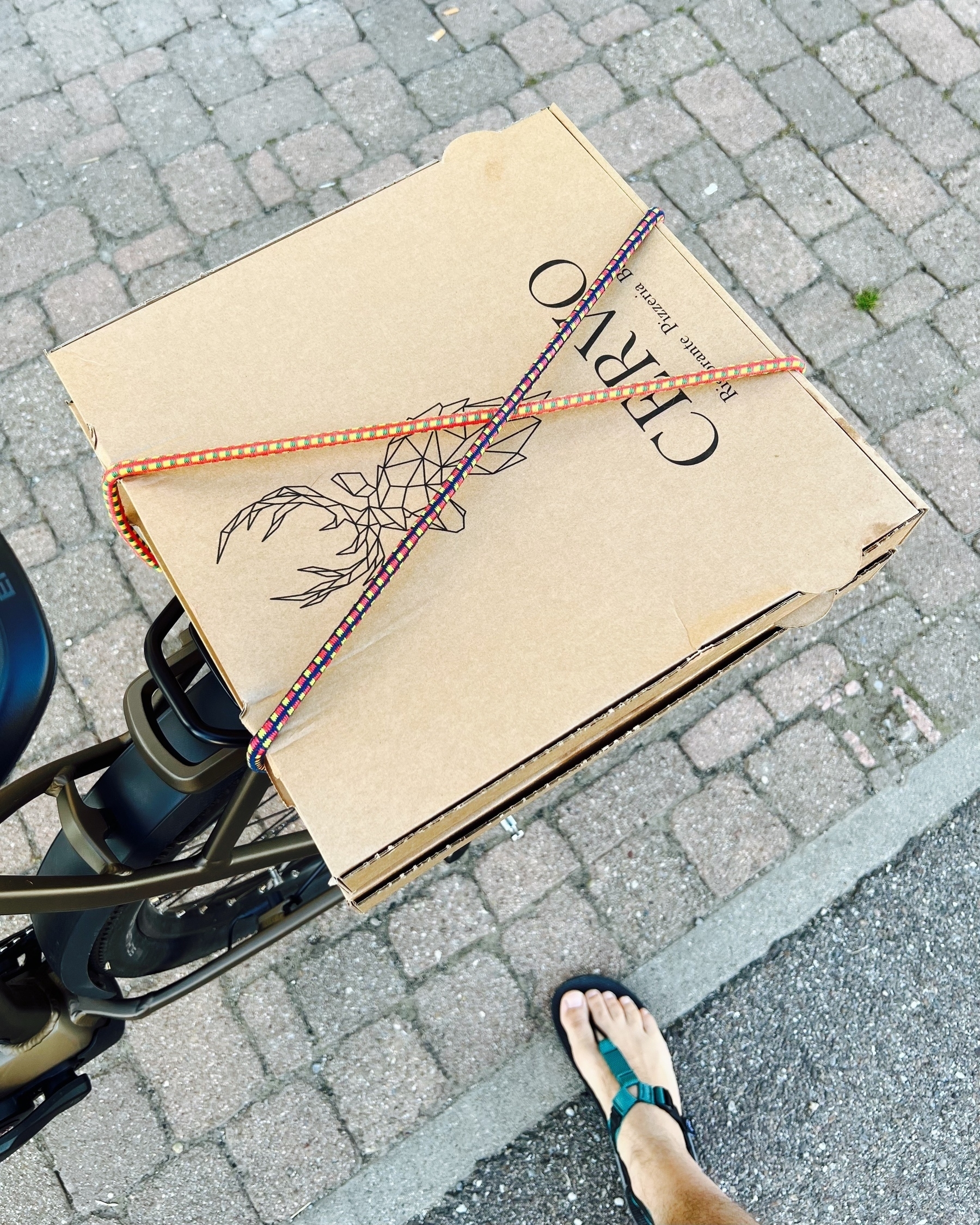 Two brown pizza boxes with a very simple design strapped to the rear rack of a bronze e-bike with elastic bands. at the bottom of the frame a right foot in sandals is partly visible.