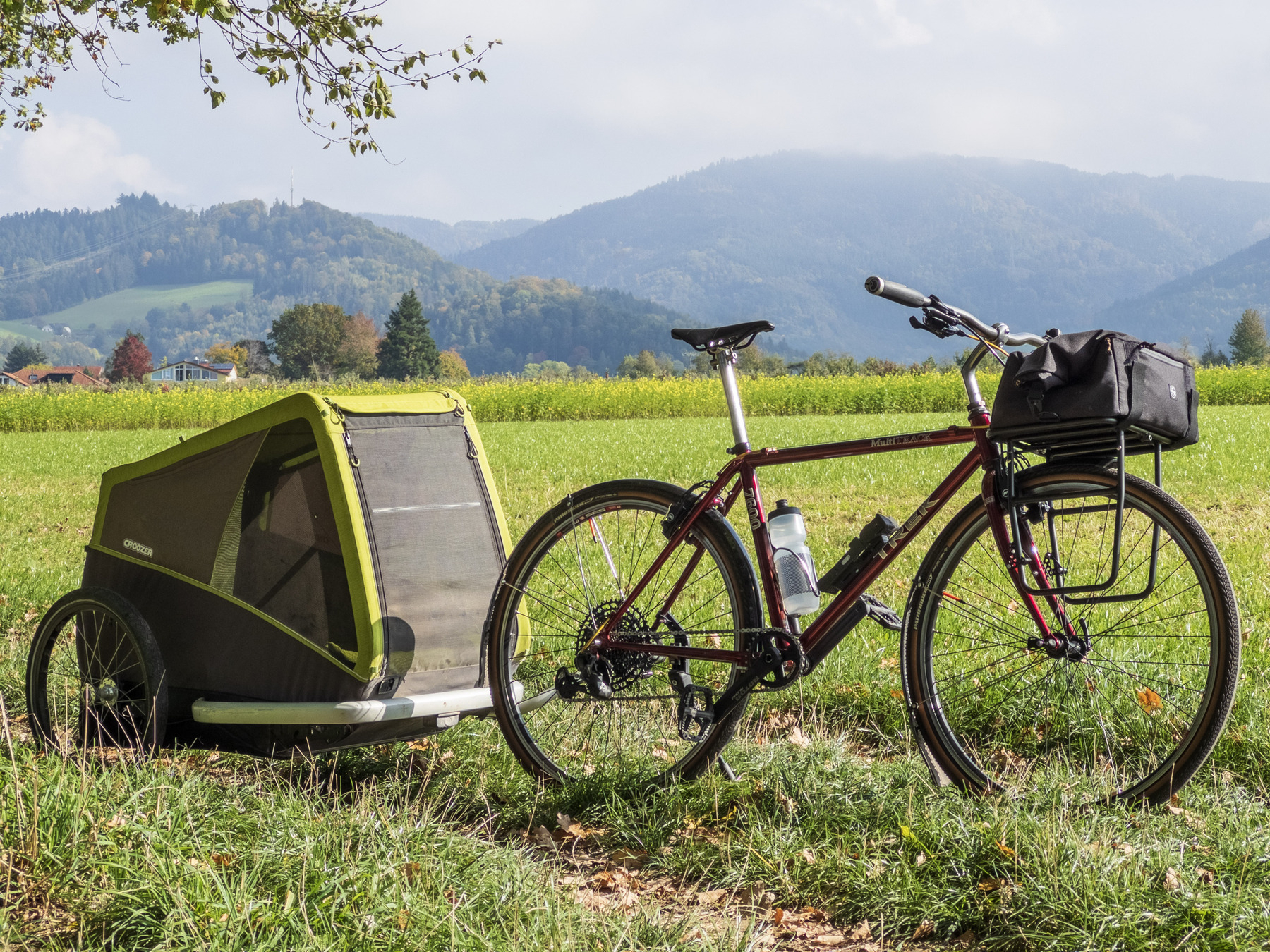 Retro-mod of a 1996 Trek MultiTrack rigid trekking MTB with a Croozer dog trailer attached. The bike and trailer are standing on a grassy path alongside a green field, in front of a slightly hazy mountain vista.