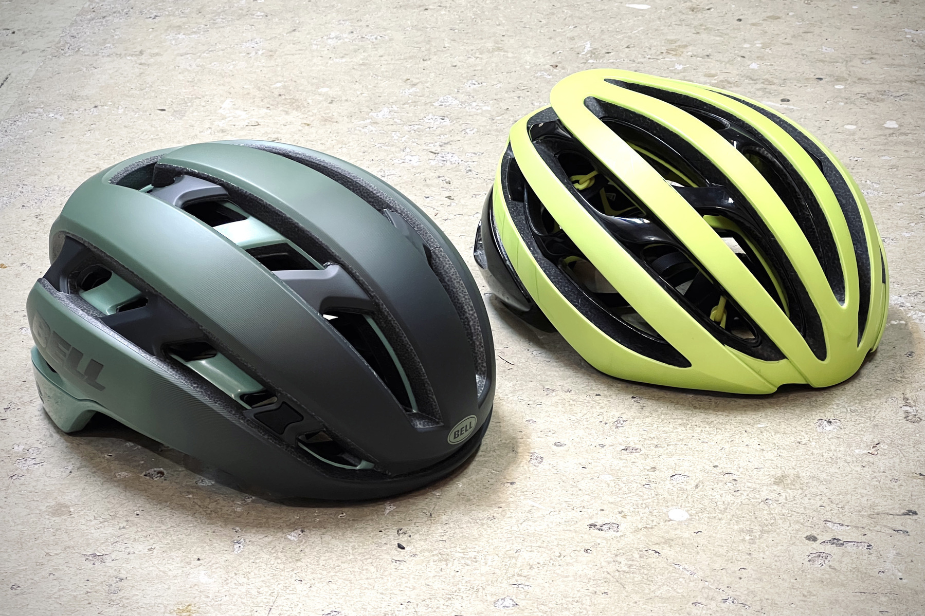 Two cycling helmets laying on a sealed concrete floor. A dark grey and green helmet in the foreground and on the left and a neon yellow helmet diagonally behind it and on the right. Both helmets face the viewer at an angle. The green helmet has straighter lines and a cleaner design overall while the neon yellow helmet has a more fluid, organic design with bigger vents.