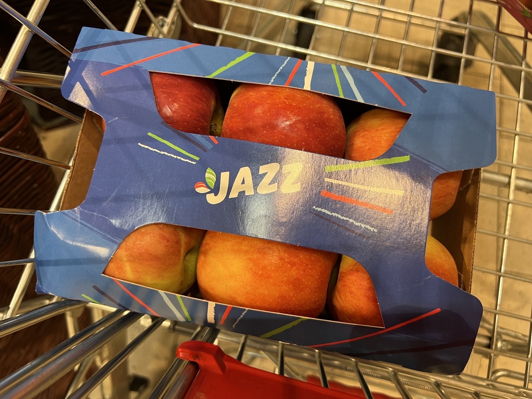 A carton sixpack of apples of the type „Jazz“ in a metal shopping cart photographed close up and from the top.