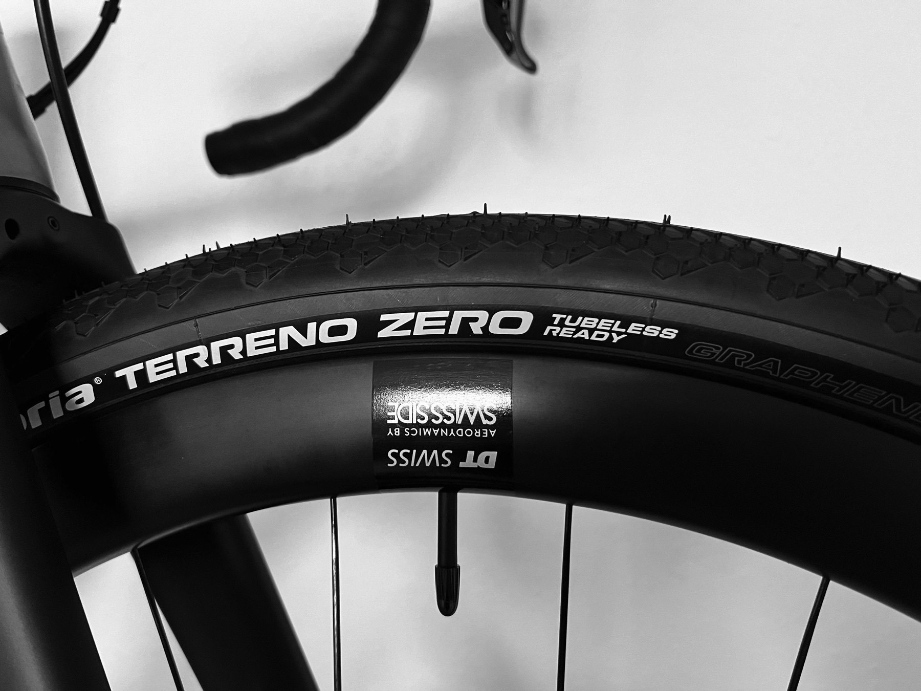 Upper fifth of a mixed surface tyre with a black semi-slick tread and grey sidewalls. The tyre is mounted on a high-flanged carbon rim by DT Swiss. The tube valve is visible. Outside of the center of the image, parts of the carbon for and the left drop and brake/shift lever of the handlebars is visible.