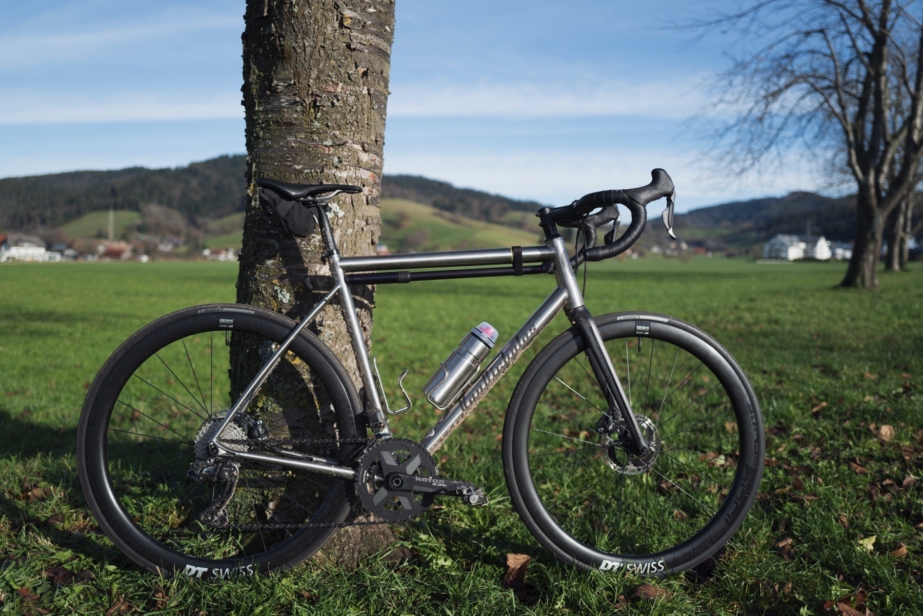 Unpainted titanium road bike with a black carbon fork leaning against a tree on a meadow. The bike is dirty and covered in mud. The blurry background shows hills with grass and forests.