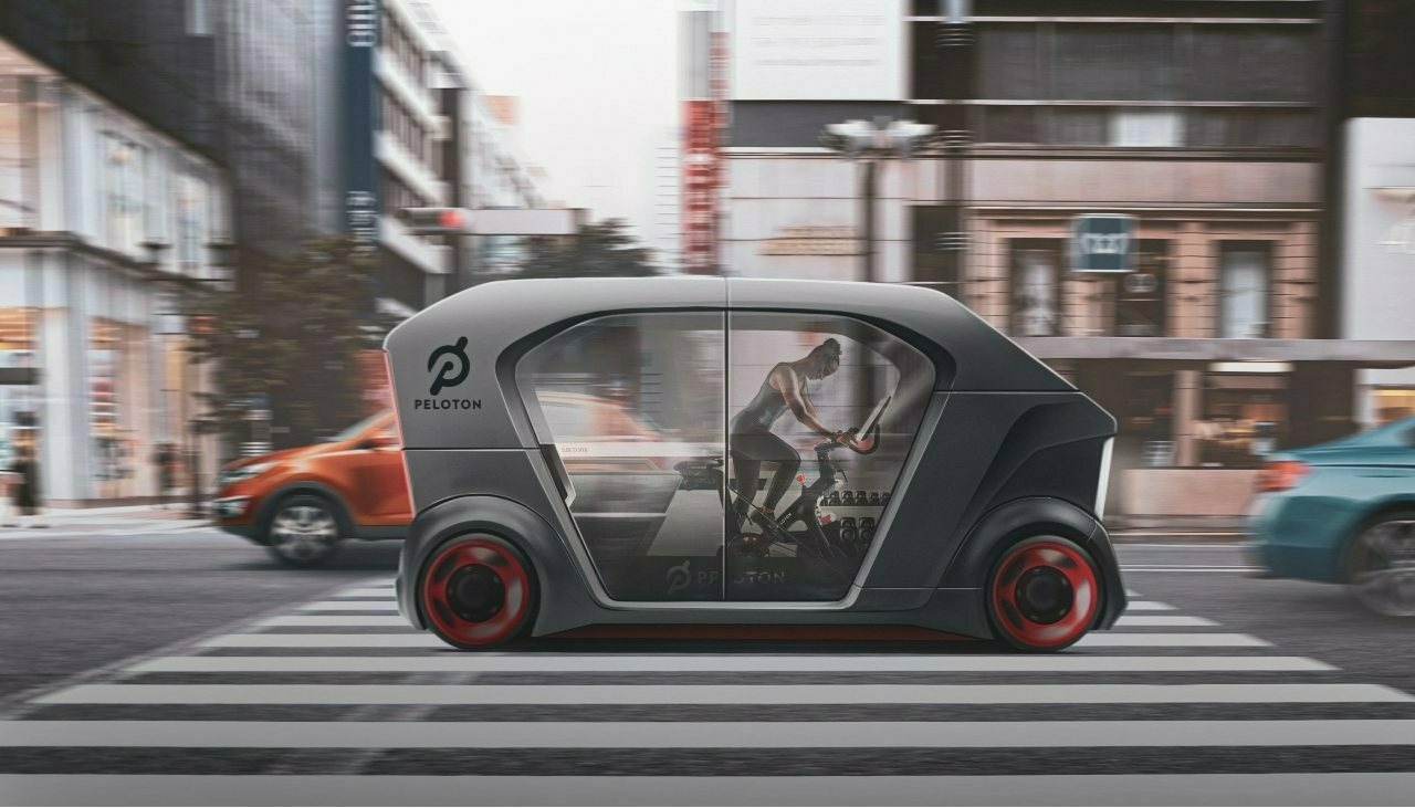 Design mock-up of an autonomous vehicle driving across an intersection in a city, housing a small gym and specifically a Peloton indoor training bicycle. A female person can bee seen working out on the Peloton bicycle inside the vehicle.