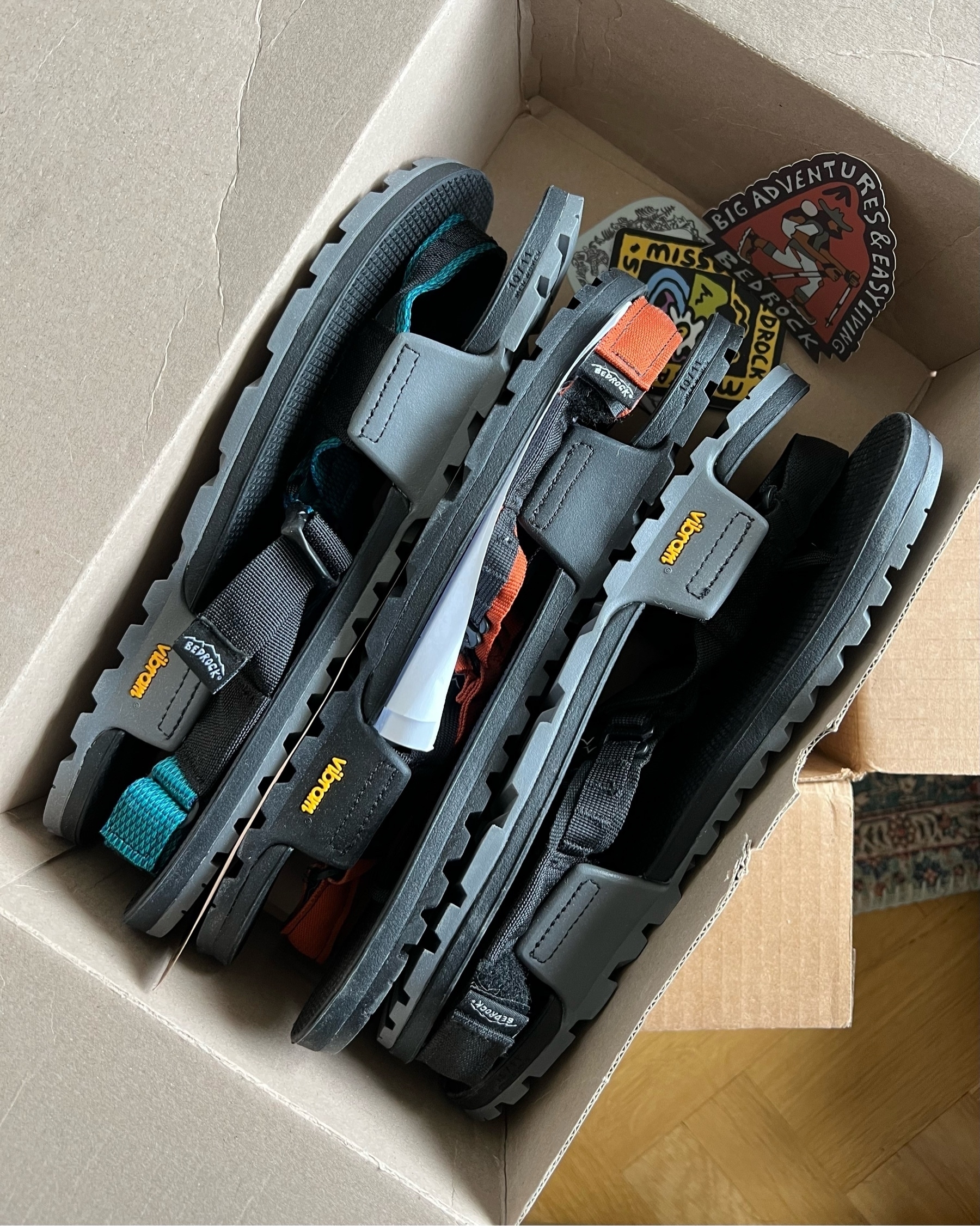 Three pairs of Bedrock Cairn sandals tightly packed in a cardbord box. Three colourful stickers laying in one end of the box.