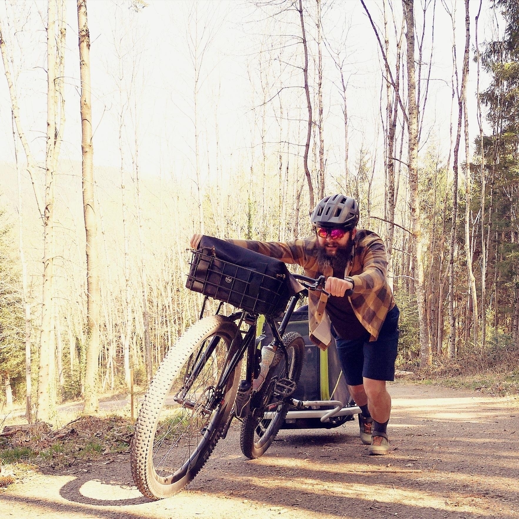 Man pushing a mountain bike with an attached dog trailer holding a dog up a very steep incline. The man is leaning over an into the task intently because od the strain.