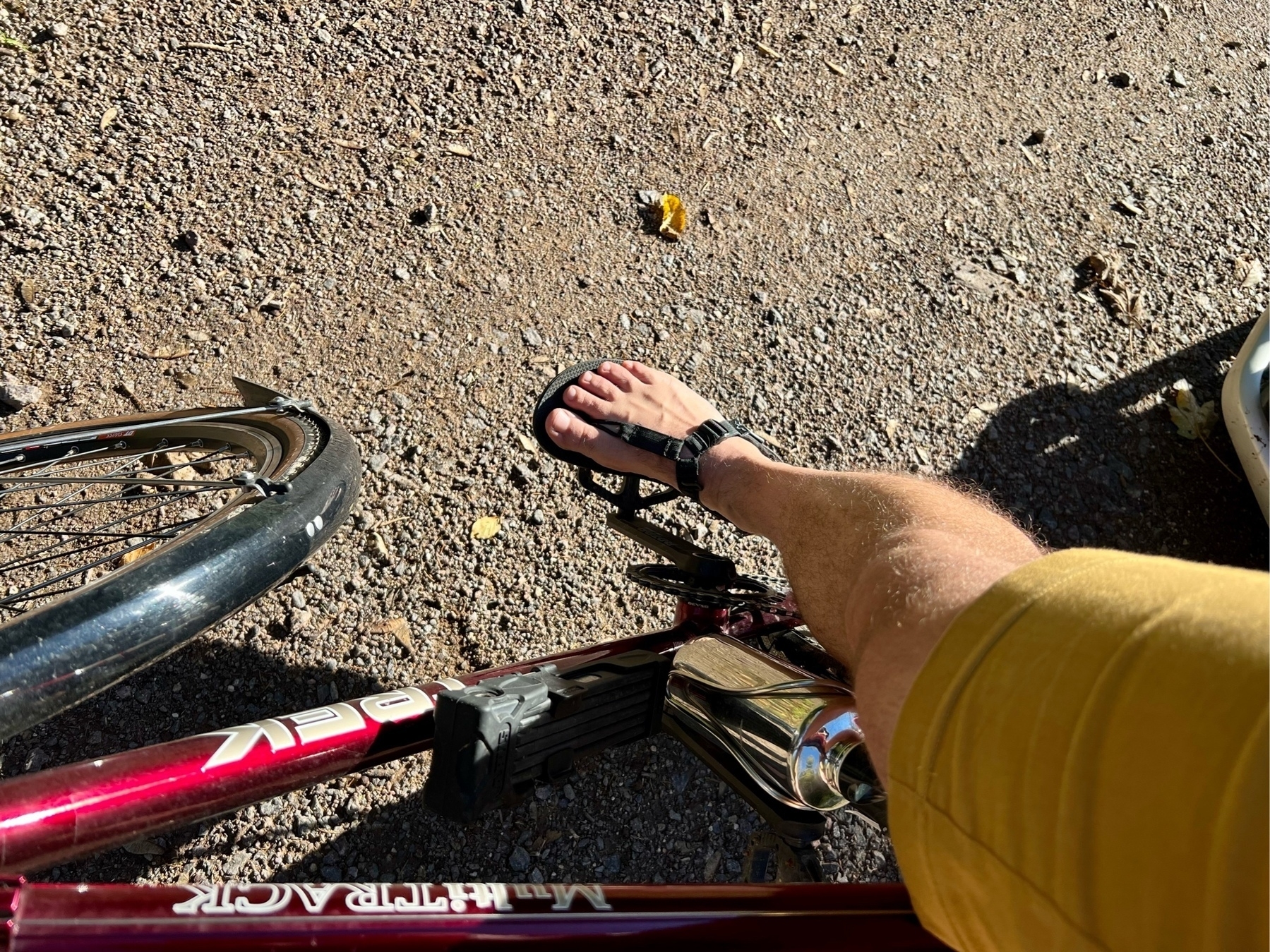 Downward look at an outstrechted leg standing on a pedal over a red bike frame. The person is wearing yellow shorts and black sandals.